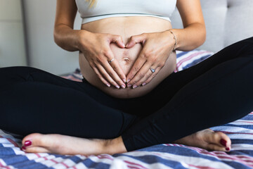 Pregnant woman making heart shape with hands on her bump while sitting on the bed