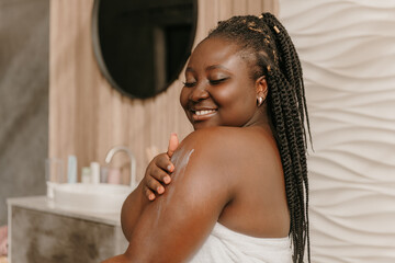 Beautiful plus size African woman covered in towel applying hydrating cream on shoulder in bathroom - obrazy, fototapety, plakaty