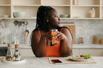 Bored plus size African woman holding glass with juice while having lunch at home