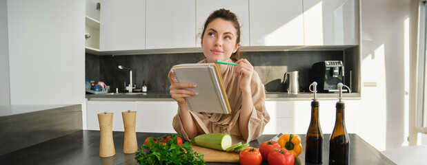 Portrait of woman checking recipe notes in notebook, standing in kitchen with vegetables, cooking...