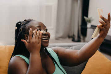 Happy plus size African woman applying eye patches and looking at the mirror while relaxing at home