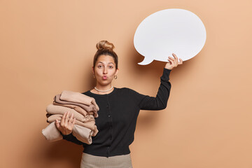 Young pretty European woman with hair bun clutching folded attire and blank speech bubble looks surprisingly at camera keeps lips folded wears casual black jumper isolated over brown background