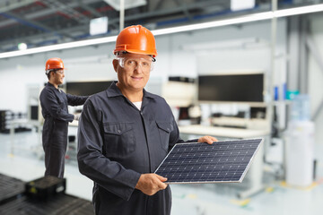Worker in a uniform wearing helmet and goggles and holding a solar panel in a factory