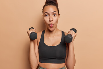Waist up shot of surprised Latin womn lifts dumbbells trains to have muscles has sporty body dressed in black cropped top isolated on brown background leads healthy lifestyle. Working out with weights