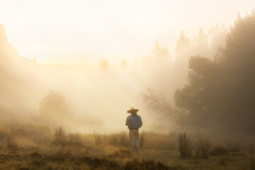 Woman in nature in meadow sunset with mist golden light, back with straw hat, subject in the foreground not in focus - obrazy, fototapety, plakaty