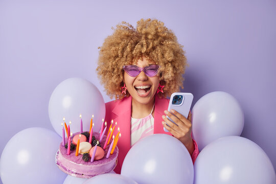 Overjoyed positive curly haired woman holds taty festive cakke and smartphone receives congratulations on her anniversary poses among balloons enjoys party time isolated over purple background