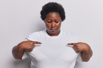 Horizontal shot of plump dark skinned woman points two fingers at mockup space of tshirt wears casual t shirt poses against white background suggests to write your logo here. Clothes and promotion - obrazy, fototapety, plakaty