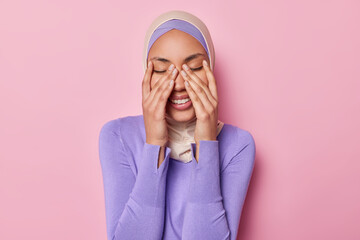 Horizontal shot of positive Muslim woman keeps eyes closed keeps hands on face smiles gladfully wears traditional hijab laughs at something funny isolated on pink background. Positive emotions concept