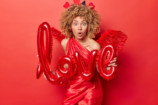 Indoor shot of young African american woman wearing costume of Cupid celebrating St. Valentine's Day organising party surprised having so many guests looking straight at camera with love sign