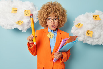 Indoor shot of shocked curly haired female teacher finds out astonished information holds big pencil and folders surrounded by sticky notes with written reminder dressed in formal clothes isolated
