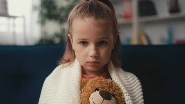 Unhappy little girl hugging teddy bear while sitting alone on couch, sadness
