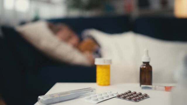 Sick little girl sleeping on sofa with teddy bear, pills and medication on table