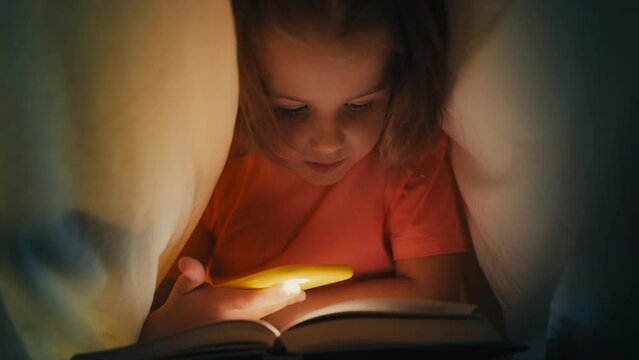 Little girl reading a book under blanket at night, using phone flashlight