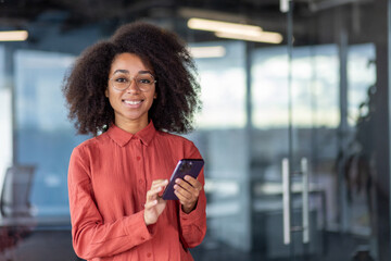 Portrait of young beautiful successful african american business woman inside office at work place inside office, female worker smiling and looking at camera, using app on smartphone