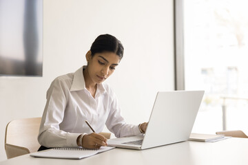 Serious pretty Indian business professional girl writing notes at laptop compute, sitting at workplace table in office, talking to client on video conference call, studying online