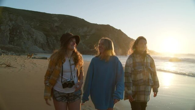 Three best friends, young women walk on empty sunset beach, laugh and smile. Concept of freedom and millennial youth generation. Happy female friends in cinematic scenery