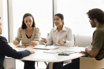 Positive motivated young Indian professional woman offering creative idea for project to listening coworkers on brainstorming meeting, sitting at table, speaking, smiling, working with team