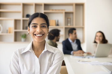 Happy young beautiful Indian businesswoman office portrait. Positive pretty business professional girl looking at camera, posing in co-working meeting room with colleagues talking behind