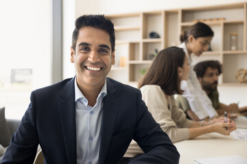 Fototapeta na wymiar Cheerful handsome mature Indian business leader man professional portrait. Happy businessman, boss, company owner, project manager sitting at meeting table with team of employees working in background