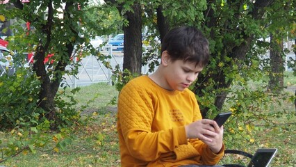 smiling happy fun teenage schoolboy boy sits in a park on a bench use mobile cell phone chat online rest relax outdoors Urban. spending time on social networks, online training, conference. video call