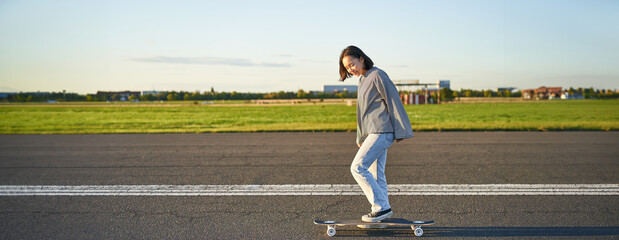 Beautiful asian skater girl riding her longboard on sunny empty road. Young woman enjoying her...