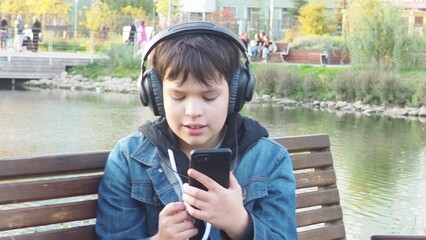 smiling happy fun teenage schoolboy boy sits in a park on a bench use mobile cell phone chat online rest relax outdoors Urban. spending time on social networks, online training, conference. video call