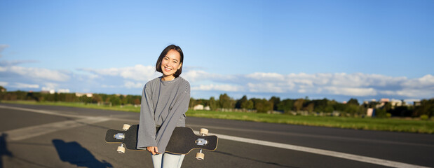 Vertical shot of skater girl posing with longboard, cruising on empty road in suburbs. Smiling...