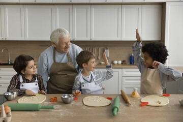 Happy multigenerational family cooking in kitchen. Three cute multiethnic boys prepare homemade cookies with help of older great-grandpa, have fun, enjoy process, dessert preparation, giving high five - obrazy, fototapety, plakaty