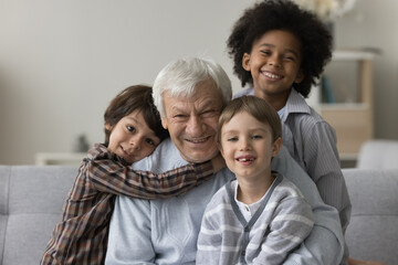 Three multiracial grandsons hug their hoary great-grandfather, smile look at camera, sitting on...