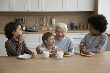 Multigenerational family, three cute great-grandchildren spend time with loving elderly great-grandpa sit in kitchen drinking milk eating delicious sweet homemade cookies, enjoy food and conversation