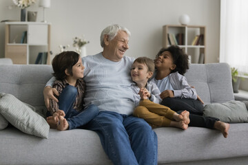 Three multiethnic adorable children boys enjoy conversation with loving hoary great-grandfather, multigenerational family spend weekend time together, having pleasant trustworthy talk in living room