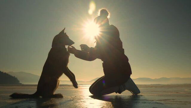 CLOSE UP, LENS FLARE, SILHOUETTE: Lady and her adorable dog practice high five on a frozen lake. They stopped to learn a new trick while ice skating across a big lake on a cold and sunny winter day.
