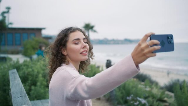 Calm relaxed young female tourist in casual elegant beige cardigan making selfie picture on front camera for social media blog or to send to friends and family in ocean view park at summer vacation 4K
