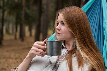 Red-haired girl lying in a hammock and drinking tea. A happy young woman resting in the autumn forest.