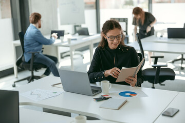 Smiling female entrepreneur holding note pad while working on laptop sitting in modern coworking