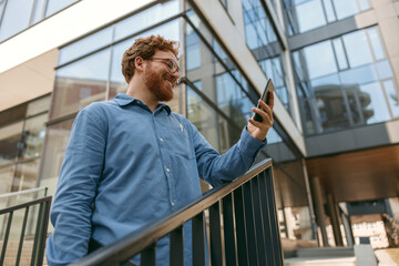 Smiling male freelancer in eyeglasses is use phone standing on coworking building background