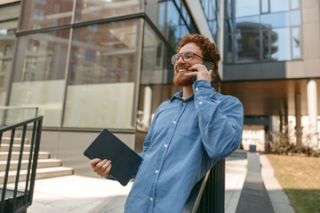 Fototapeta premium Smiling male manager in eyeglasses is talking phone standing near office building
