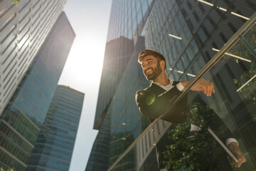 Stylish businessman is standing with laptop on office terrace and looks away. High quality photo