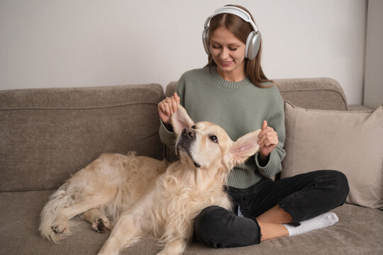 Girl sitting with her dog on the couch with headphones on. Young woman stroking her dog at home.