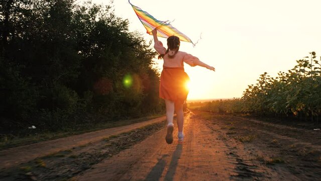 Joyful little girl with kite runs along rural road at back sunset light