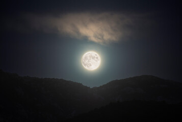 Full moon above mountains on cloudy sky, horizontal landscape of Earth satellite in full lunar phase