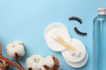 Bottle of makeup remover, cotton flowers, pads, swabs and false eyelashes on light blue background, flat lay. Space for text