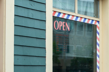 The exterior of a green wooden building with cream colored trim. The shop has an illuminated red open sign hanging in the glass window of the barbershop. There's a red, white and blue striped edging.  - Powered by Adobe