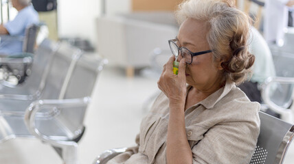 Old lady using inhalant to relief her headache waiting to see doctor at a clinic or hospital lobby