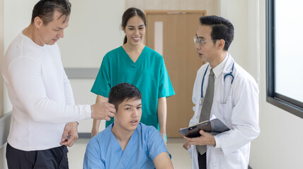 Fototapeta premium Young boy disable patient is sitting on a wheelchair under support from nurse and doctor to move around the hospital corridor.