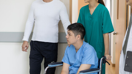 Young boy disable patient is sitting on a wheelchair under support from nurse and doctor to move around the hospital corridor.