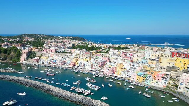 Exciting aerial truck right shot from Panoramica sulla Corricella on Procida Island (Isola di Procida) of the blue Tyrrhenian Sea in Naples' gulf with grey breakwaters, white boats, pastel buildings
