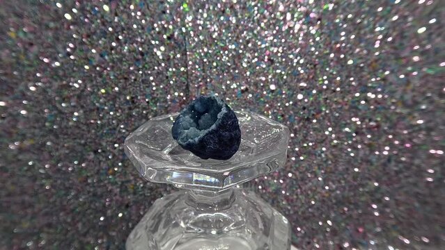 Blue geode crystal slowly rotating on a flipped glass cup on a blur silver glitter background