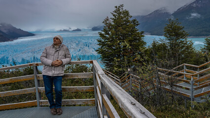 Fototapeta na wymiar A man stands on the observation deck of the glacier. He leans on the railing, looks into the distance, smiles. A mass of blue ice with peaks and cracks stretches between the mountains to the horizon.