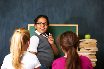 Teaching, smile and portrait of kid student by board for lesson, knowledge or education with friends. Happy, glasses and girl child talking and writing with chalk for classmates in school or academy.